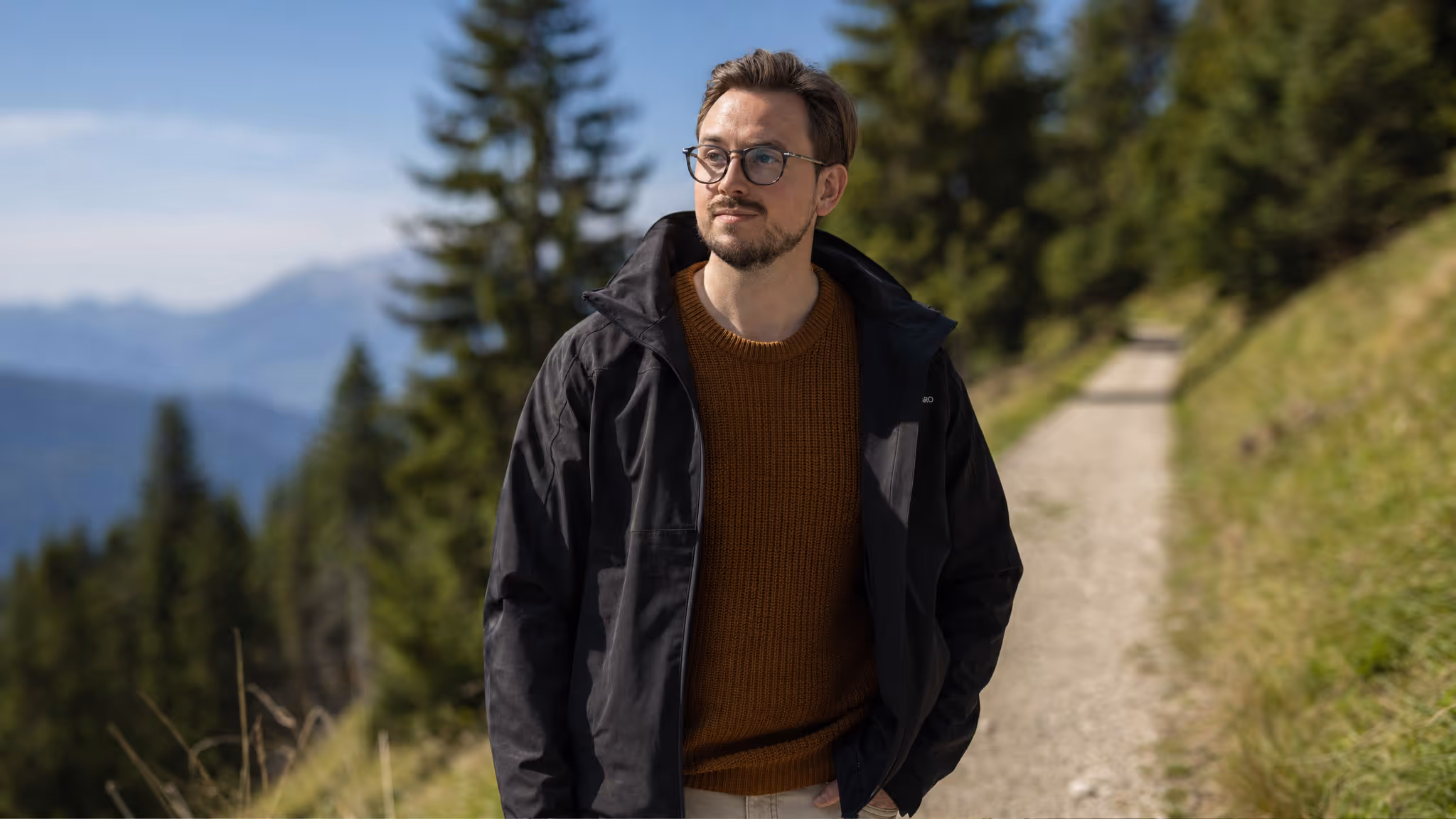 Mann mit ruhigem Blick spaziert auf einem sonnigen Bergweg durch die Natur und vermittelt Gelassenheit, Klarheit und innere Ruhe.