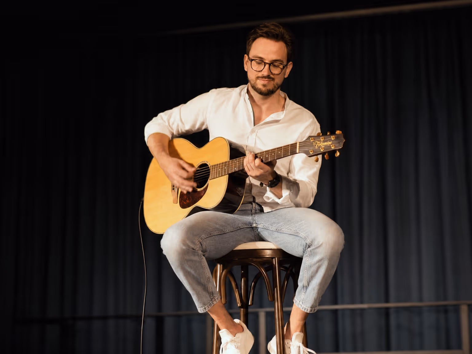 Raphael sitzt auf einem Hocker auf einer Bühne und spielt konzentriert Akustikgitarre vor dunklem Bühnenvorhang.