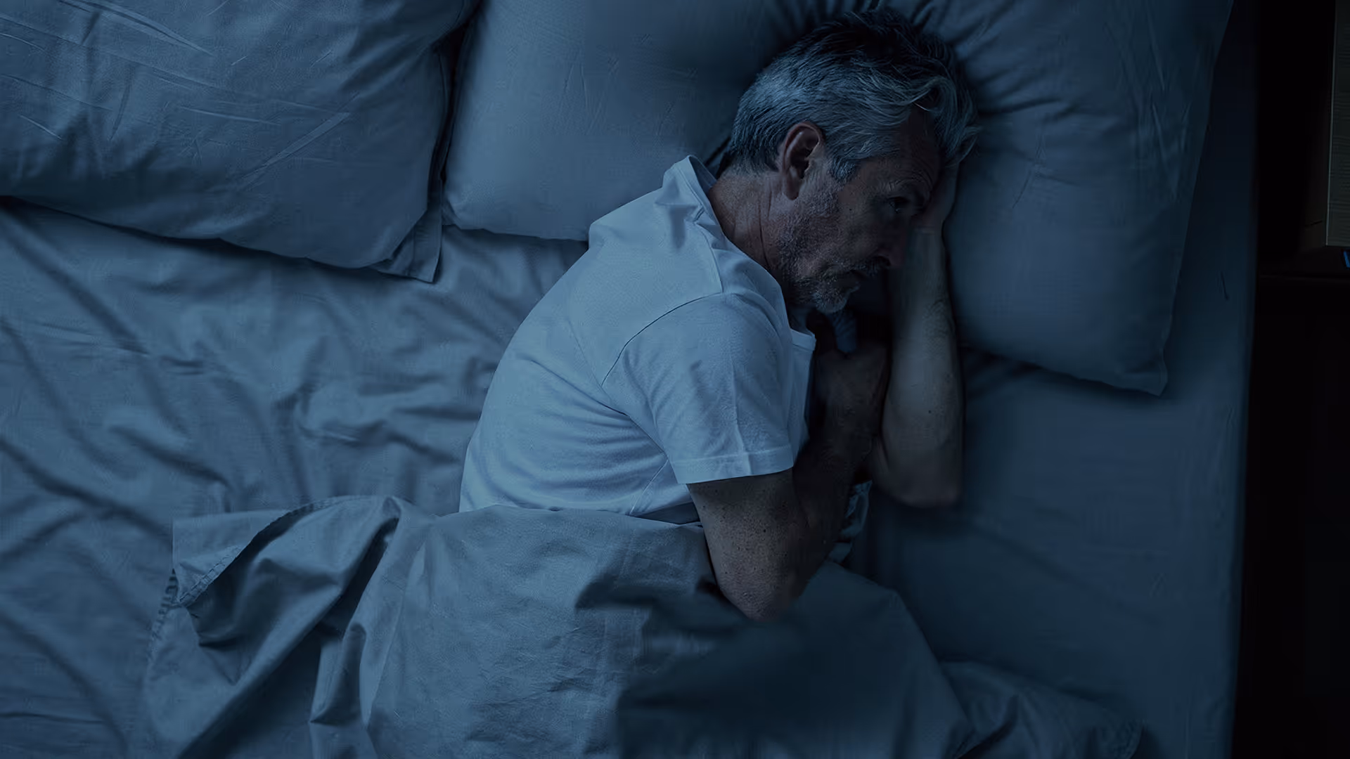 Älterer Mann liegt nachts wach im Bett und blickt angespannt zur Seite, während die dunkle Beleuchtung und seine verkrampfte Haltung Schlaflosigkeit, Grübeln und innere Unruhe vermitteln.